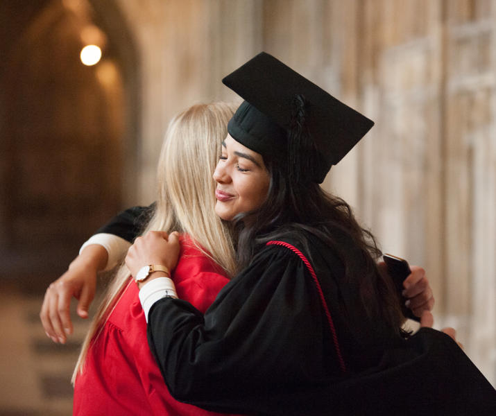 Image of a student smiling in a graduation robe and cap hugging another person